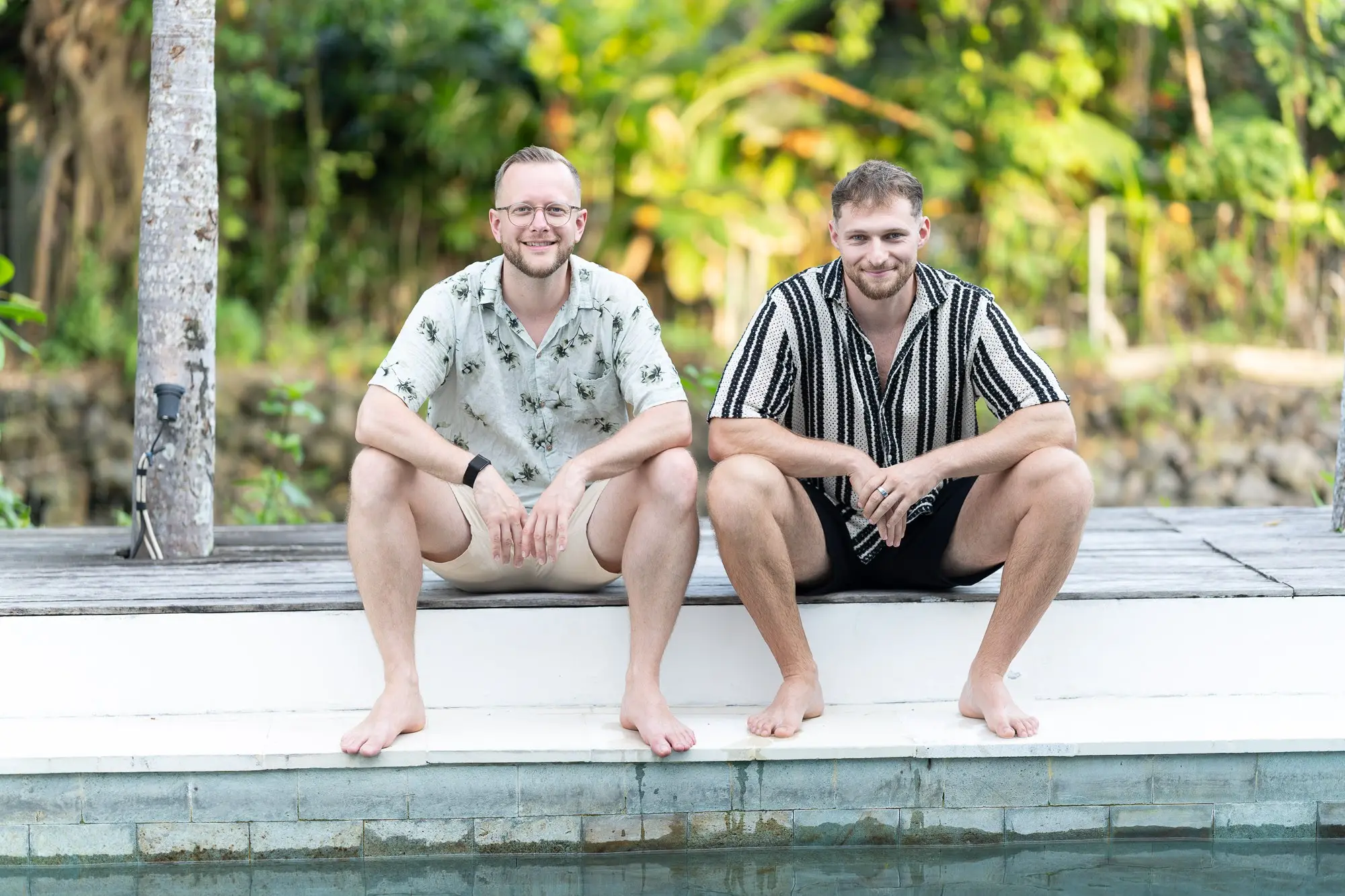 Tim und Felix sitzen lachend nebeneinander am Poolrand auf Bali
