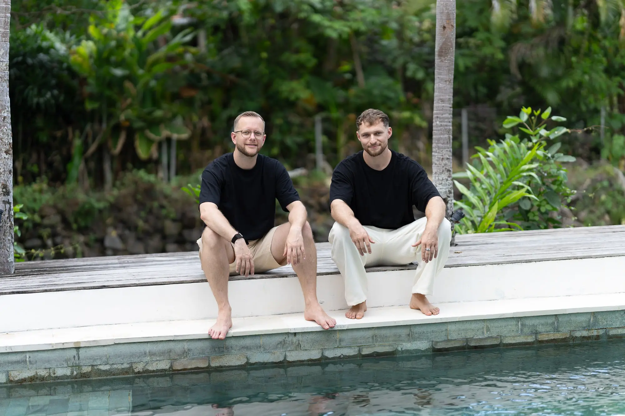 Tim und Felix sitzen barfuß am Poolrand vor tropischem Grün
