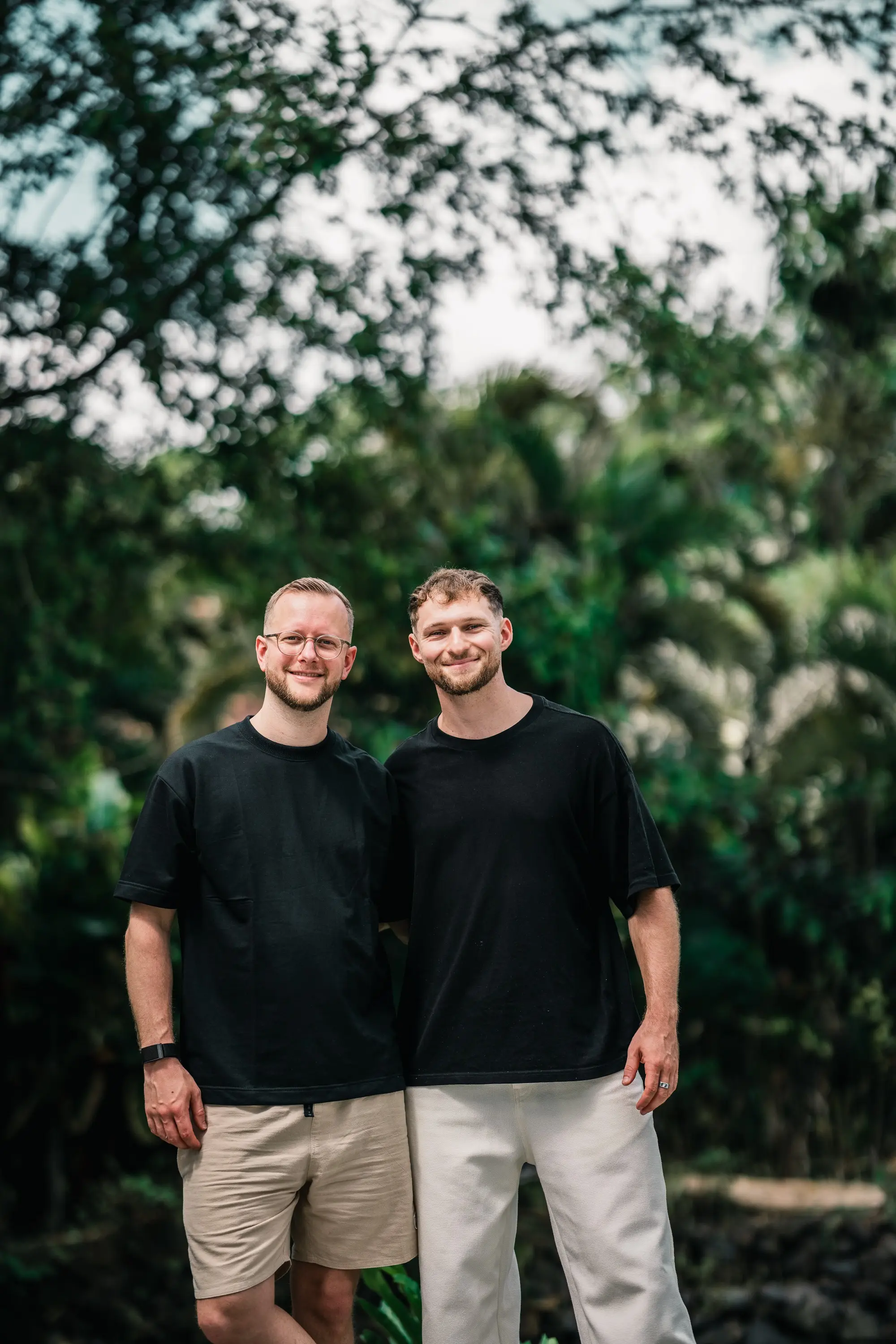 Tim und Felix stehen lächelnd nebeneinander vor tropischem Grün