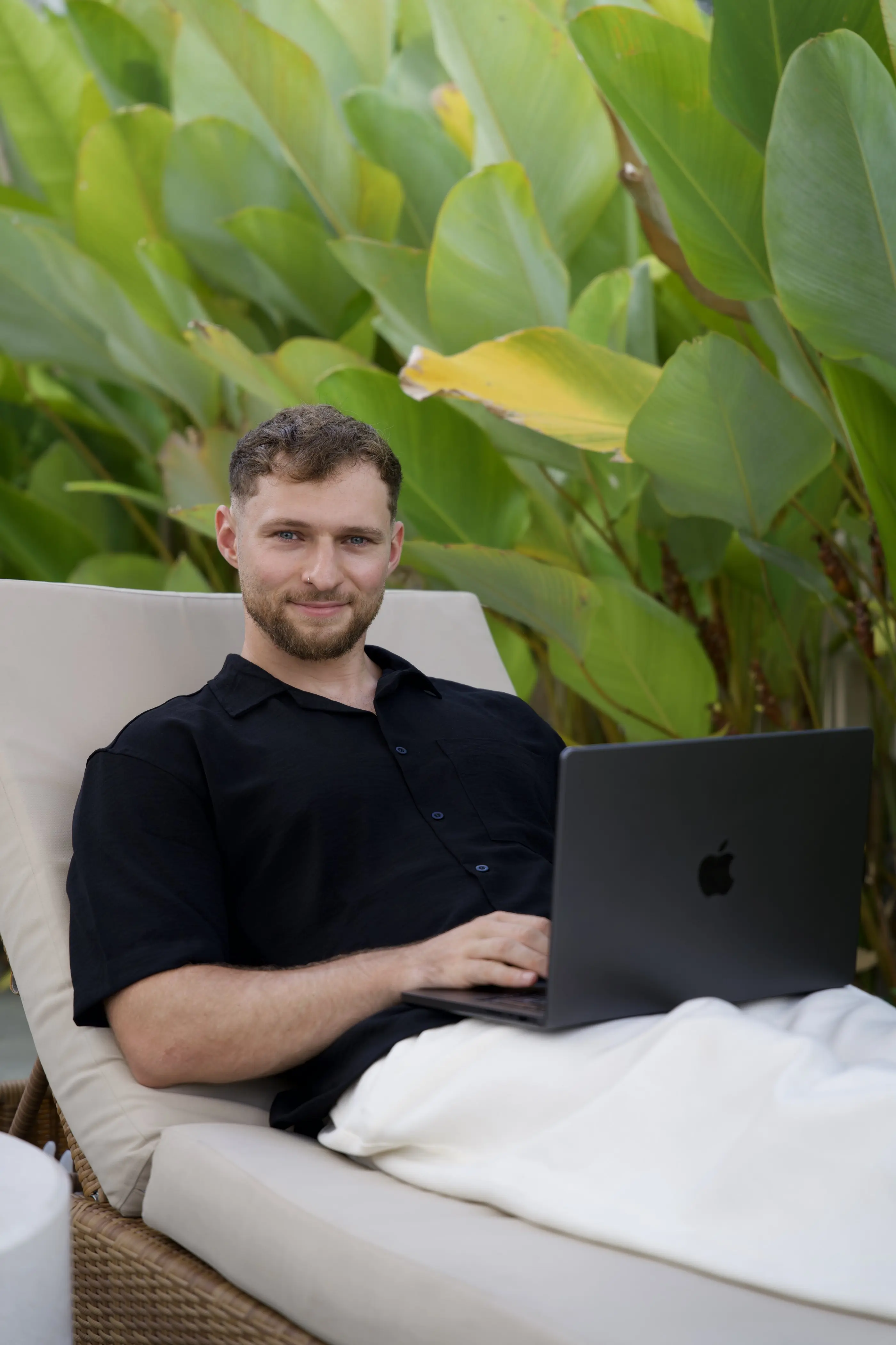 Felix sitzt mit Laptop auf einer Liege zwischen tropischen Pflanzen
