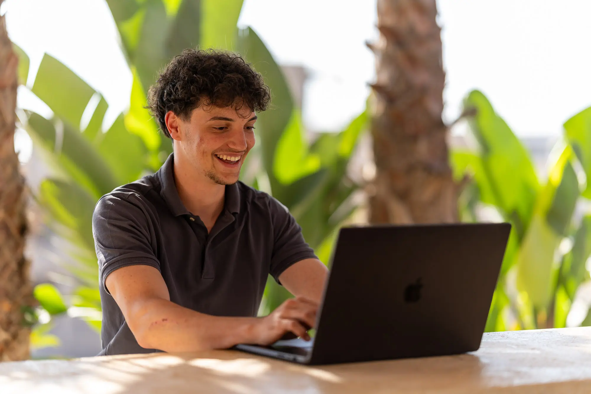 Alec lacht beim Arbeiten am Laptop vor Palmen