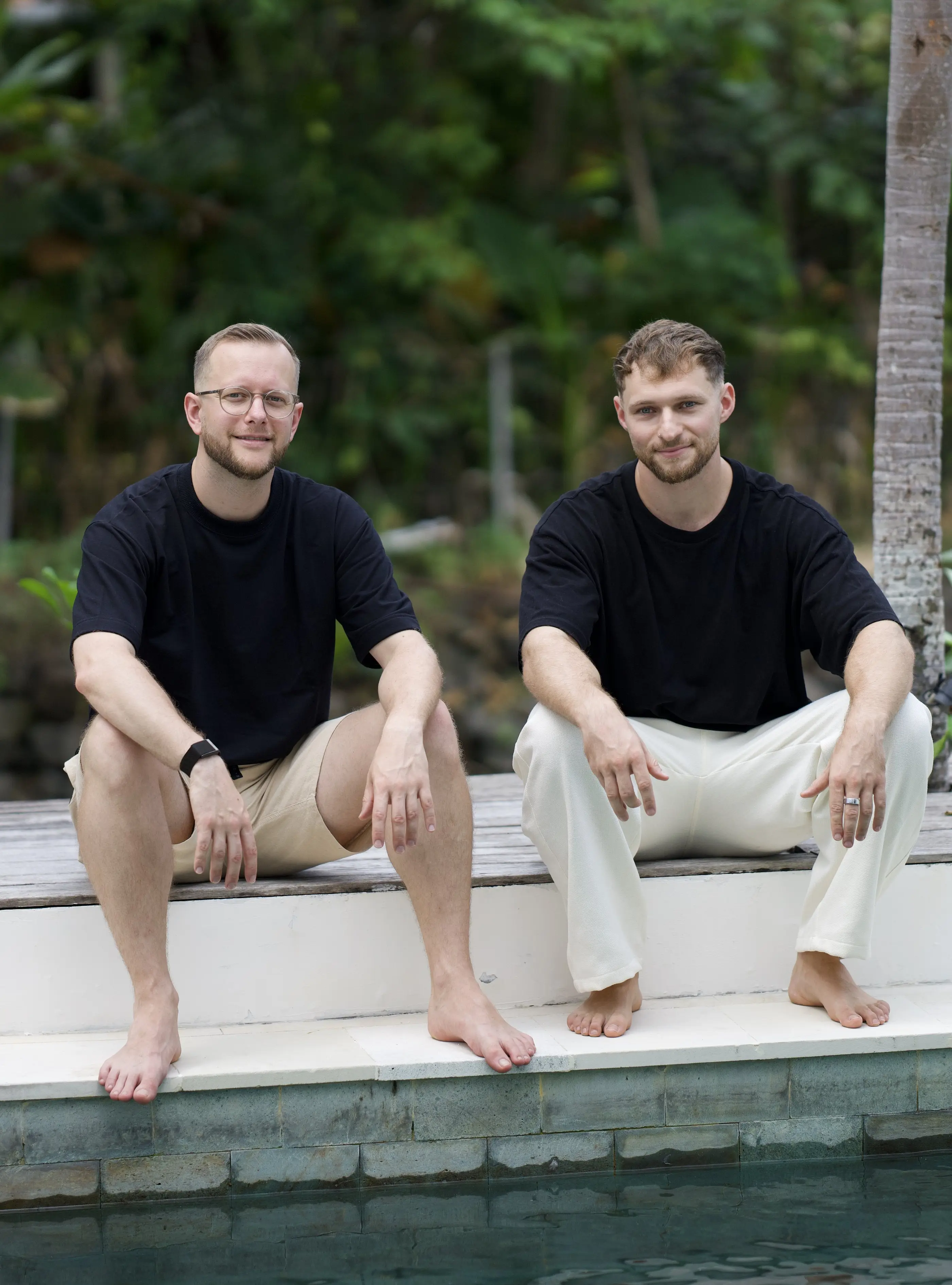 Tim und Felix sitzen barfuß am Poolrand vor tropischem Grün