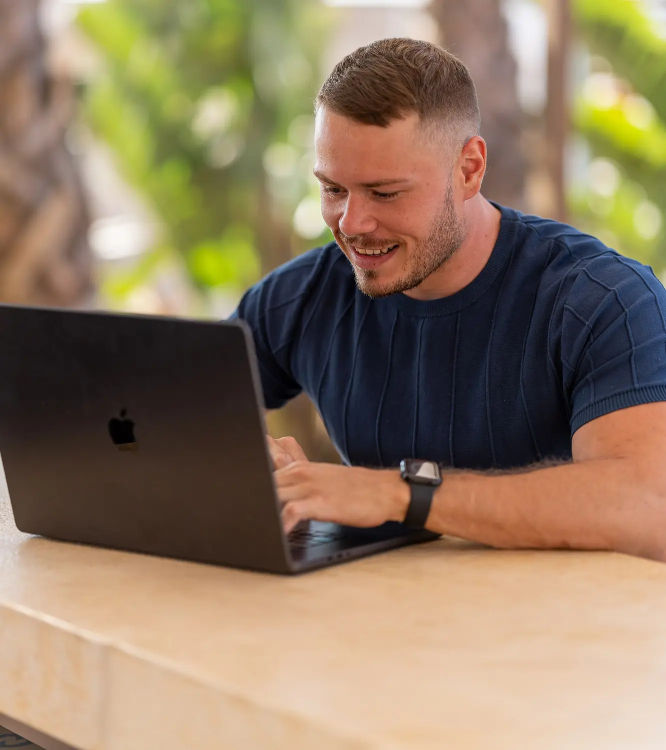 Mario arbeitet lächelnd am Laptop vor Palmen