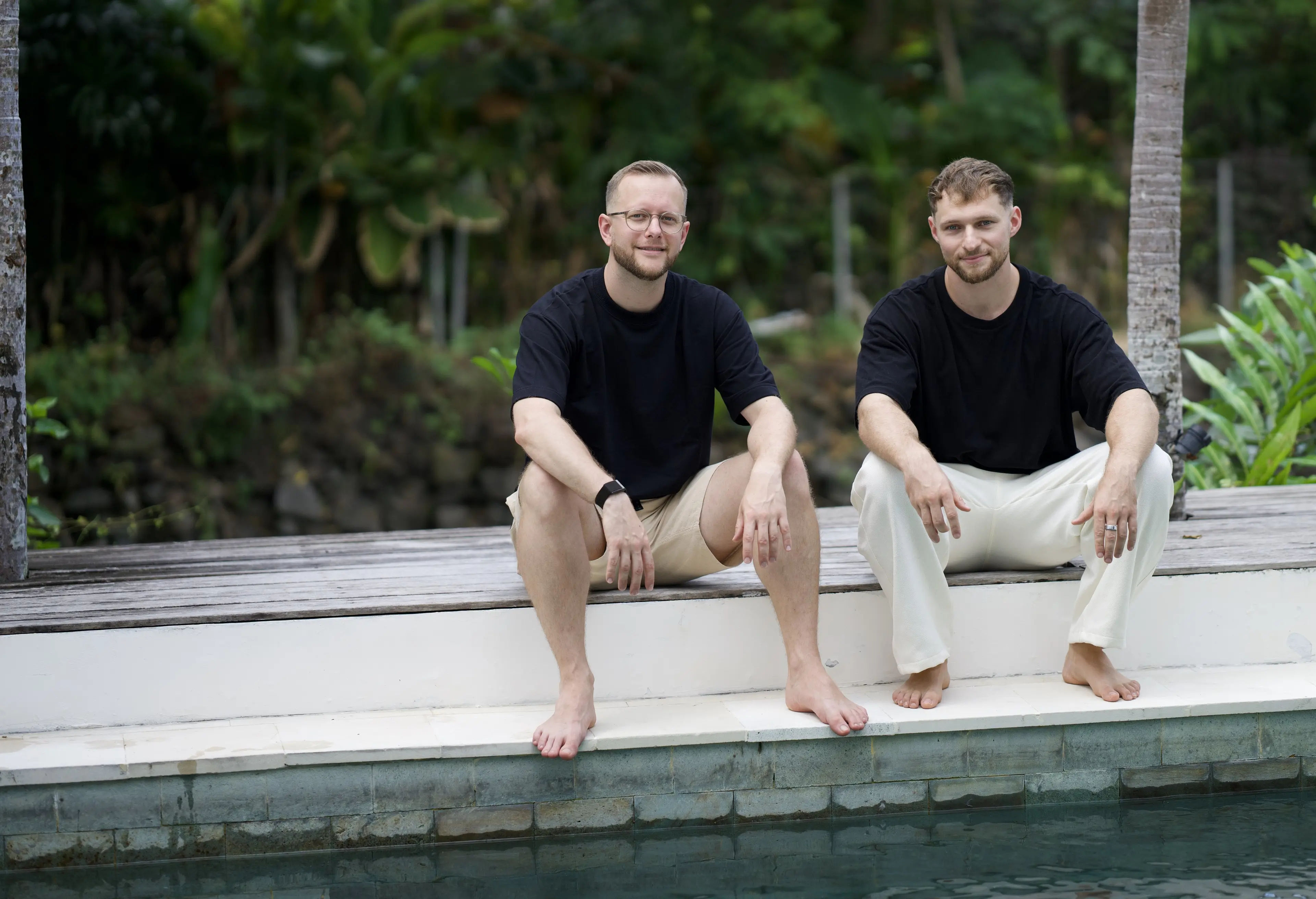 Tim und Felix sitzen barfuß am Poolrand vor tropischem Grün